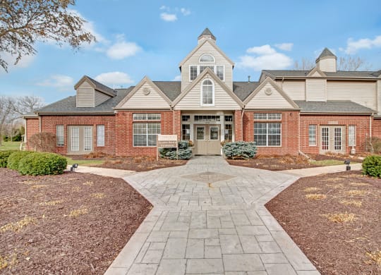 St. James Place Apartments. Brick building with peaked roofs and dormer windows, set against a blue sky. Central entrance with a pathway leading through landscaped grounds.