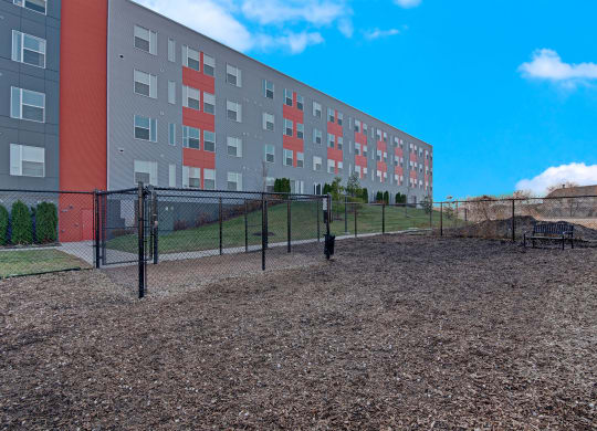 Fenced dog park with mulch ground in a modern residential area. Four-story gray and red apartment building in the background, under a bright blue sky at Stitchweld Apartments.