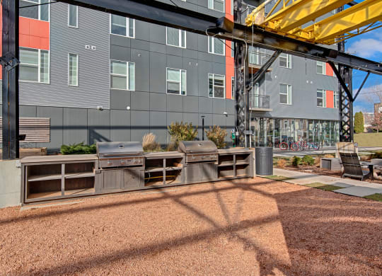 Outdoor area with two metal grills under a modern structure, adjacent to a gray building. Bicycles and lounge chairs are visible. Bright, sunny day at Stitchweld Apartments.