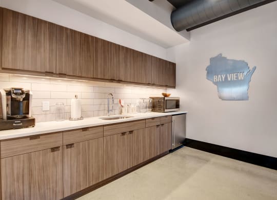 Modern kitchen with wooden cabinets, white countertops, and a stainless steel sink. A sign shaped like Wisconsin says "eat well" on the wall, creating a welcoming vibe at Stitchweld Apartments.