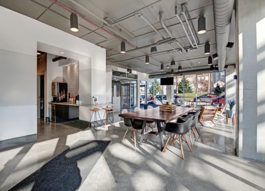 Modern cafe interior with large windows and natural light. Features a long wooden table with black chairs, a coffee station, and minimalist decor at Stitchweld Apartments.