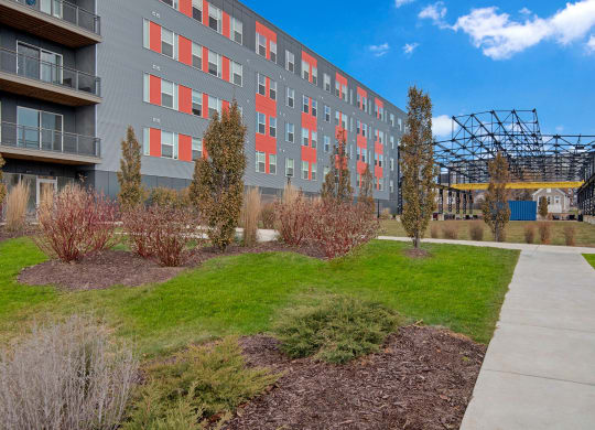 Modern apartment building with gray and red panels, surrounded by landscaped greenery. A pathway and metal structure are visible under a clear blue sky at Stitchweld Apartments.