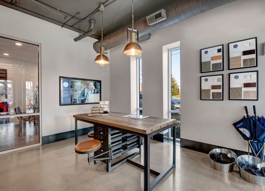 Modern office lobby with industrial decor. A wooden table and stool set is centered under two pendant lights. Sample boards and umbrellas line the walls, creating a welcoming atmosphere at Stitchweld Apartments.