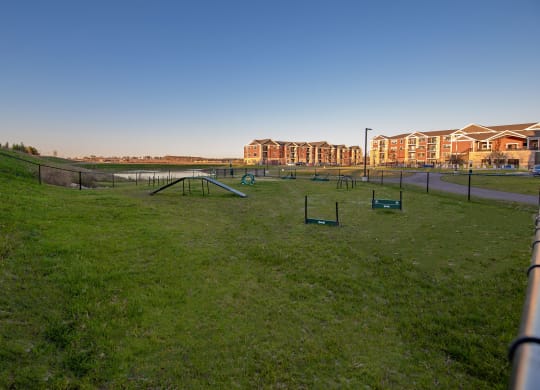 A grassy dog park with agility equipment, fenced and bordered by The Park Apartments, a row of red-brick apartments in the background.