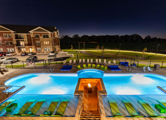 Night view of a brightly lit swimming pool with green lounge chairs and a central fire pit at the Park Apartments. An apartment building and parked cars are in the background.