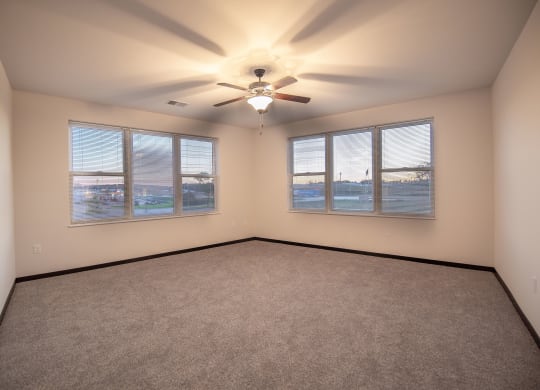 Spacious empty room at the Park Apartments with beige walls and carpet, ceiling fan, and large windows with blinds showing a sunset sky, creating a serene atmosphere.