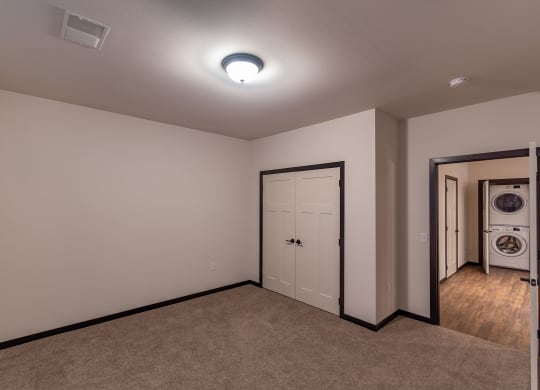 An empty room with beige walls and carpet flooring features a closed double door and an open door leading to a laundry area with a stacked washer and dryer at The Park Apartments.