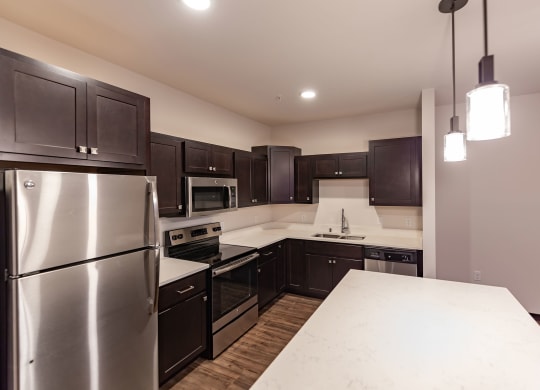 Modern kitchen at the Park Apartments with dark wood cabinets, stainless steel appliances, and white countertops. Pendant lights hang over the island, creating a sleek, cozy atmosphere.