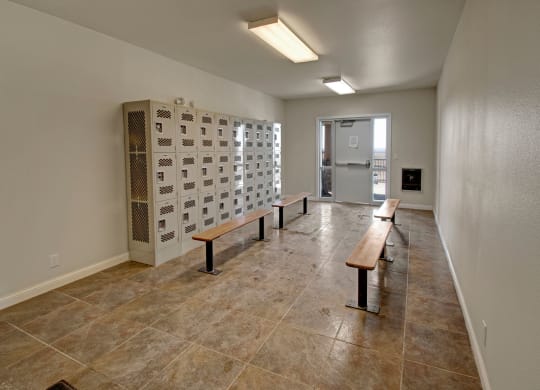 A clean, empty mail room here at Confluence at the Harvest Hills Apartment Homes with beige floor tiles, metal lockers lining the left wall, and wooden benches in the middle. Overhead fluorescent lighting.