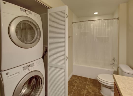 Compact laundry and bathroom space here at Confluence at the Harvest Hills Apartment Homes with stacked washer and dryer on the left, white bathtub with a curtain, toilet, and tiled floor. Clean and functional.