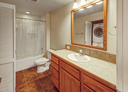 Compact bathroom here at Confluence at the Harvest Hills Apartment Homes with wooden cabinets below a sink and mirror. White toilet and shower with curtain on the left, stacked washer and dryer visible. Warm lighting.