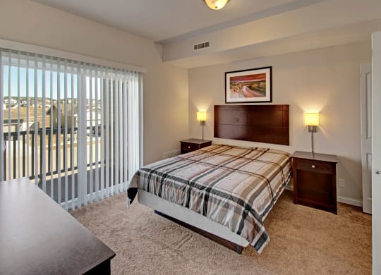 A cozy bedroom here at Confluence at the Harvest Hills Apartment Homes with a plaid-patterned bed, flanked by two nightstands and wall lamps. Large window with vertical blinds offers natural light.