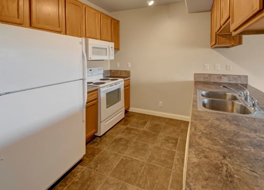 Compact kitchen here at Confluence at the Harvest Hills Apartment Homes with wooden cabinets, white appliances including fridge, stove, and microwave. Spacious brown tiled countertop with double sink. Neutral tones convey simplicity.
