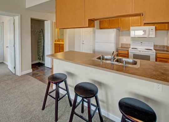 A cozy kitchen here at Confluence at the Harvest Hills Apartment Homes with wooden cabinets, a white stove, fridge, and microwave. A bar counter with three stools faces the sink, conveying a welcoming vibe.