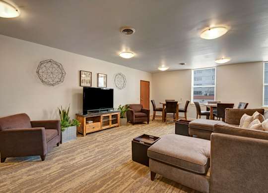 A welcoming resident lounge here at Latitude featuring warm brown seating, a large TV console, and dining tables by the windows, creating a comfortable and inviting space for relaxing or gathering.