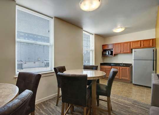 A bright dining area here at Latitude featuring round tables, dark brown upholstered chairs, and large sunlit windows, creating a warm, welcoming space connected to a cozy kitchen with wood cabinetry.