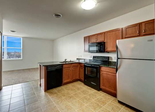 A bright kitchen here at Latitude featuring warm wood cabinetry, black appliances, and tiled flooring, opening to a spacious, inviting living area with soft carpet and a large sunlit window.
