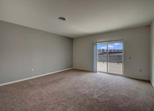 A bright living room here at Latitude featuring soft neutral carpeting, spacious layout, and a large sliding glass door opening to a private balcony, creating an airy, inviting, and comfortable atmosphere.
