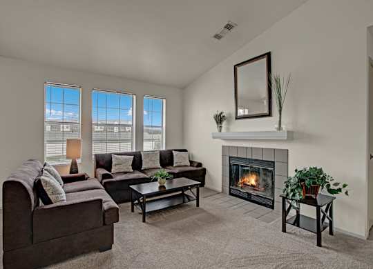 A cozy living room here at St. James Place with soft beige carpet, cream walls, and a warm corner fireplace. A comfortable seating area sits beside tall windows that bring in natural light, creating a bright and inviting atmosphere.