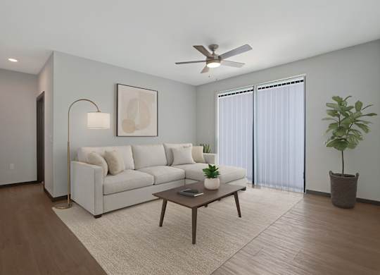 A bright living room here at The Lafayette featuring a cream sectional sofa, warm wood coffee table, light rug, sliding glass doors with vertical blinds, ceiling fan, and a tall green plant in the corner.