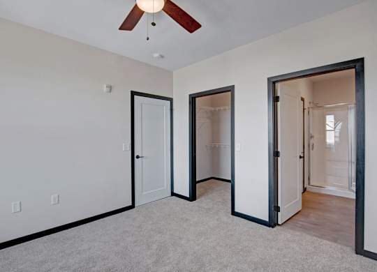 A bright bedroom here at The Lafayette with soft beige carpet, light walls, a ceiling fan, and dark trim. A walk-in closet and private bathroom with a glass shower add convenience and comfort.