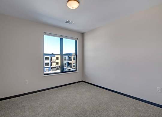 A bright bedroom here at The Lafayette with soft beige carpet, light walls, and dark trim. A large window brings in natural light and offers a pleasant view of the surrounding community buildings.