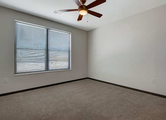 A comfortable bedroom here at The Lafayette with soft beige carpet, light walls, and dark trim. A large window with blinds brings in natural light, and a ceiling fan adds comfort and airflow.