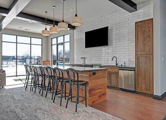 A stylish community lounge here at The Lafayette with a large kitchen island, bar seating, and warm wood cabinets. A subway tile backsplash, pendant lighting, and large windows create a bright, welcoming gathering space.