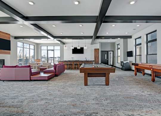 A spacious community lounge here at The Lafayette with a pool table, shuffleboard, and comfortable seating. A large kitchen island, bar seating, and bright windows create a lively, welcoming space for entertaining.