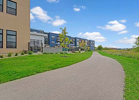 A scenic walking path here at The Lafayette with lush green landscaping and modern apartment buildings. A nearby patio area and open green space create a peaceful, inviting outdoor setting.