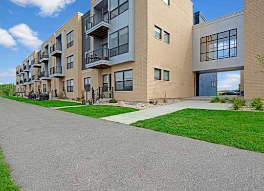 A modern exterior here at The Lafayette with private balconies, large windows, and light brick siding. A paved walking path and landscaped greenery create a clean, welcoming community setting.