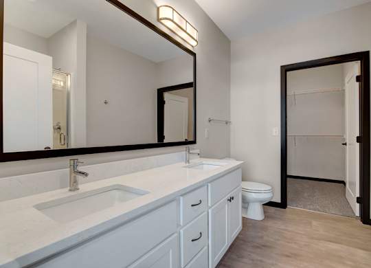 A bright bathroom here at The Lafayette featuring a wide double-sink vanity with white cabinetry, a large mirror with modern lighting, and an open doorway leading to a spacious walk-in closet.