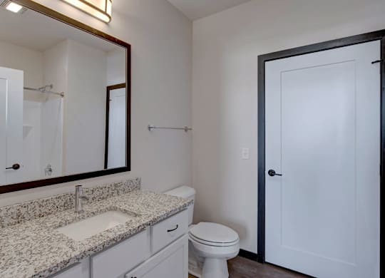 A clean bathroom here at The Lafayette featuring a bright white vanity with speckled granite countertop, a large framed mirror with modern lighting, and a sleek white door creating a fresh, inviting look.