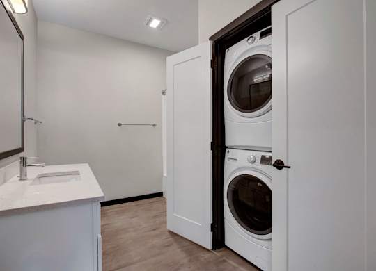A bright bathroom here at The Lafayette featuring a sleek white vanity with modern countertop, a large framed mirror, and a convenient in-unit stacked washer and dryer, creating a clean and functional space.
