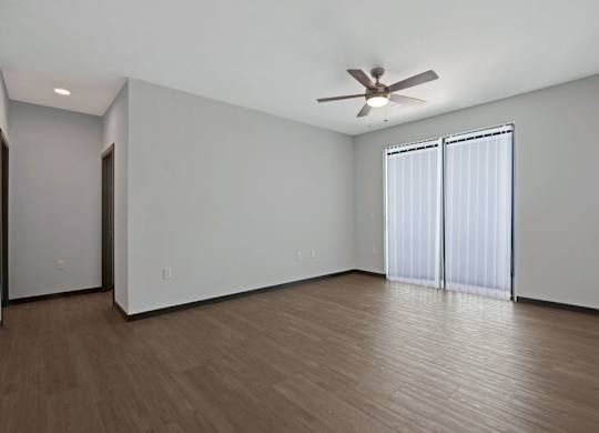 A spacious living room here at The Lafayette featuring warm wood-style flooring, soft natural light from sliding glass doors with vertical blinds, and a modern ceiling fan, creating an open and inviting atmosphere.