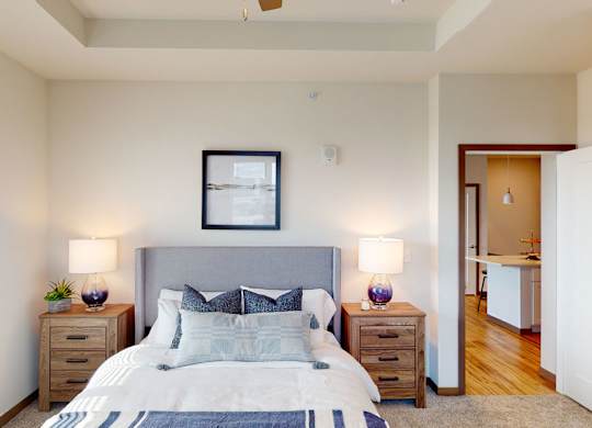 A comfortable bedroom here at Van Buren East with soft carpeting and neutral tones. A large window brings in natural light, while a cozy bed and bedside lighting create a warm, inviting atmosphere.