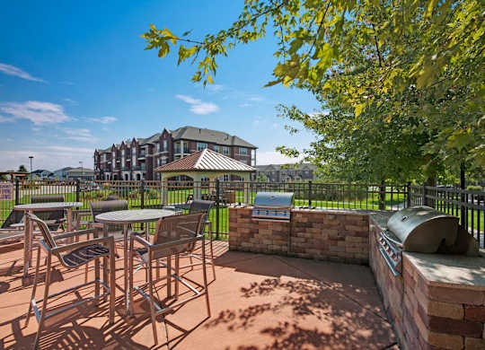 A sunny outdoor grilling patio here at Van Buren Place with built-in barbecue stations and comfortable table seating. A nearby gazebo, green lawn, and surrounding apartments create a welcoming, resort-style atmosphere.