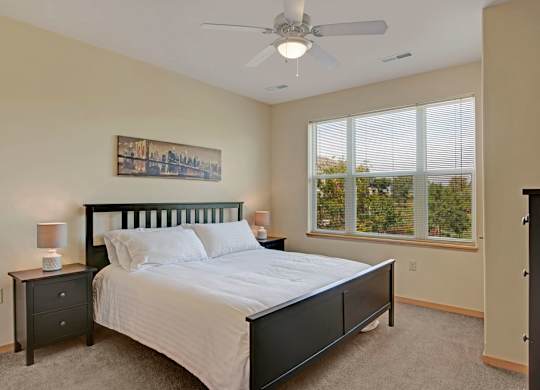 A bedroom here at Van Buren Place featuring a dark wood bed frame, soft carpet flooring, large bright windows with peaceful views, and a clean, cozy layout that feels warm and inviting.