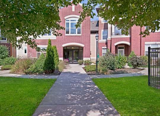 A charming building entrance here at Van Buren Place with a welcoming doorway and landscaped greenery. A shaded sidewalk, mature trees, and manicured lawn create a peaceful, inviting community setting.