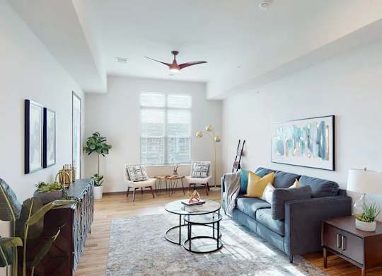 A living room here at Van Buren West featuring a deep blue sofa with yellow and teal accent pillows, round glass coffee table, light area rug, and warm wood flooring, creating a bright, airy, and inviting space with modern finishes and natural light.