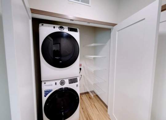 A laundry closet here at Van Buren West featuring a full-size stacked white washer and dryer, built-in white wire shelving, and warm wood-style flooring for a clean, convenient, and space-saving setup.