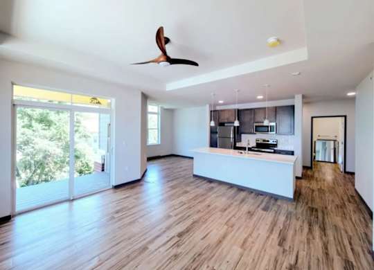 A living room here at Van Buren West featuring warm wood-style flooring, a modern kitchen island with pendant lighting, stainless steel appliances, and large sliding glass doors that fill the open layout with bright natural light.