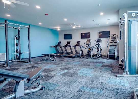 A fitness center here at Van Buren West featuring a bold teal accent wall, treadmills and stationary bikes, a cable machine, free weights, and large windows that create a bright, energetic, and well-equipped workout space.