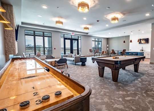 A resident lounge here at Van Buren West featuring a shuffleboard table, pool table, arcade games, and comfortable seating, with warm lighting and large windows that create a spacious, inviting, and lively gathering space.
