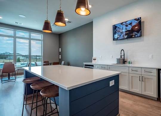 A modern resident lounge kitchenette here at Van Buren West featuring a navy island with white quartz countertops, three wood stools, warm pendant lighting, a wall-mounted TV, sleek cabinetry, and a beverage fridge, all styled in a bright, inviting layout.