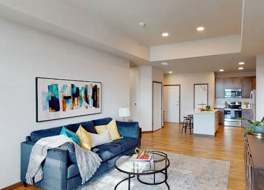 A living room here at Van Buren West featuring a deep blue sofa with yellow and teal accent pillows, round glass coffee table, light area rug, and warm wood flooring, creating a bright, open, and inviting space that flows seamlessly into the modern kitchen.