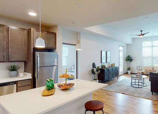A kitchen and living area here at Van Buren West featuring warm wood cabinetry, stainless steel appliances, white island with pendant lighting, and a cozy living room with dark sofa and soft blue rug, creating a bright and welcoming open-concept layout.
