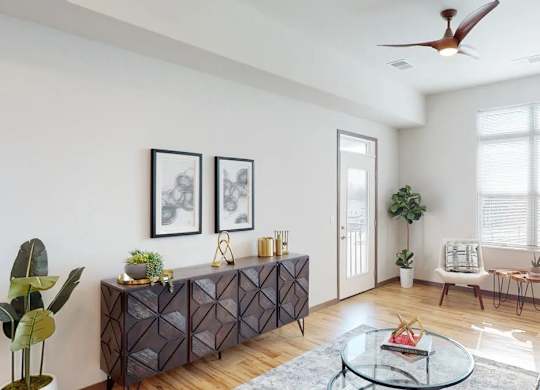 A living room here at Van Buren West featuring warm wood flooring, a dark geometric sideboard, soft gray rug, and a cozy sofa with yellow and blue accents, complemented by a bright window and inviting natural light.