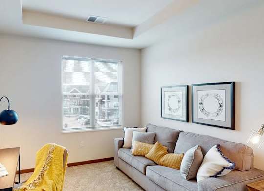 A living room here at Van Buren West featuring a light gray sofa with yellow and neutral accent pillows, soft carpet flooring, warm wood trim, and a cozy desk nook with a navy lamp and bright yellow throw for a welcoming feel.
