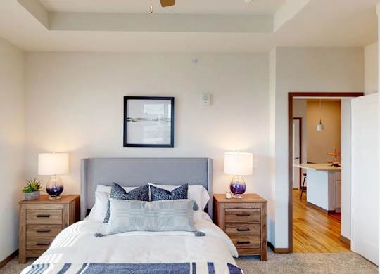 A bedroom here at Van Buren West featuring a soft gray upholstered bed, matching wood nightstands, purple glass lamps, and light neutral bedding for a bright, calm, and inviting space.